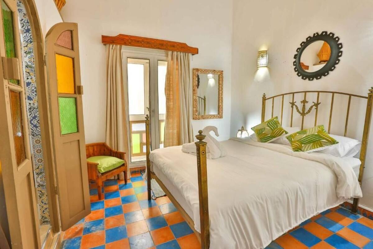 A bedroom with a checkered floor, a bed with a headboard, a chair, a door with a mirror, and a wall-mounted light fixture.
