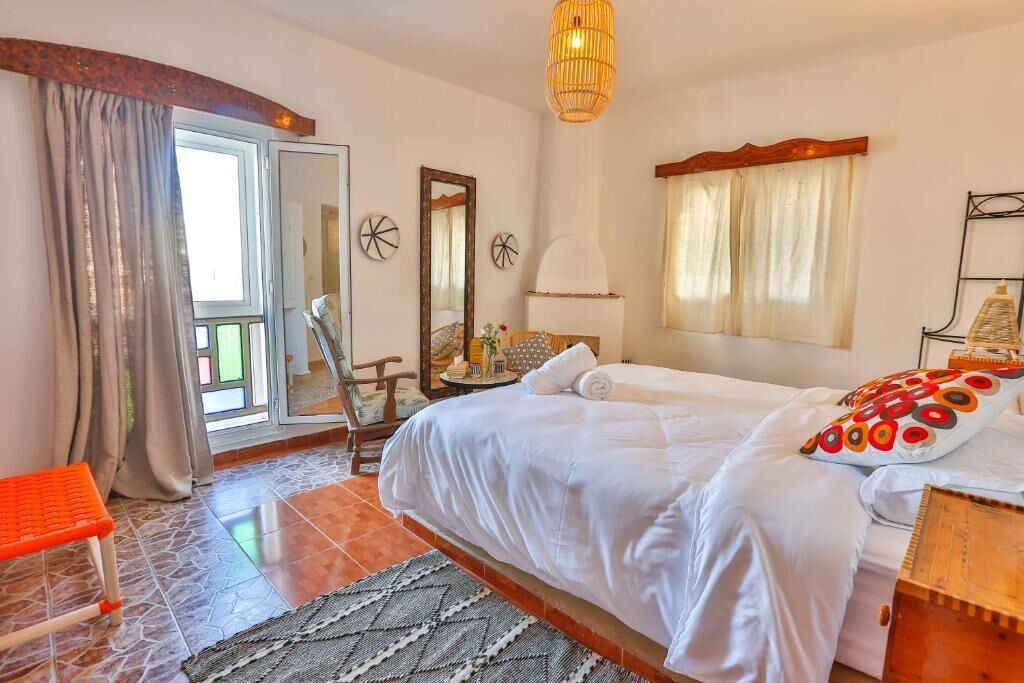 A hotel room with a bed, a chair, a table with a lamp, a window with curtains, and a door leading to a balcony.