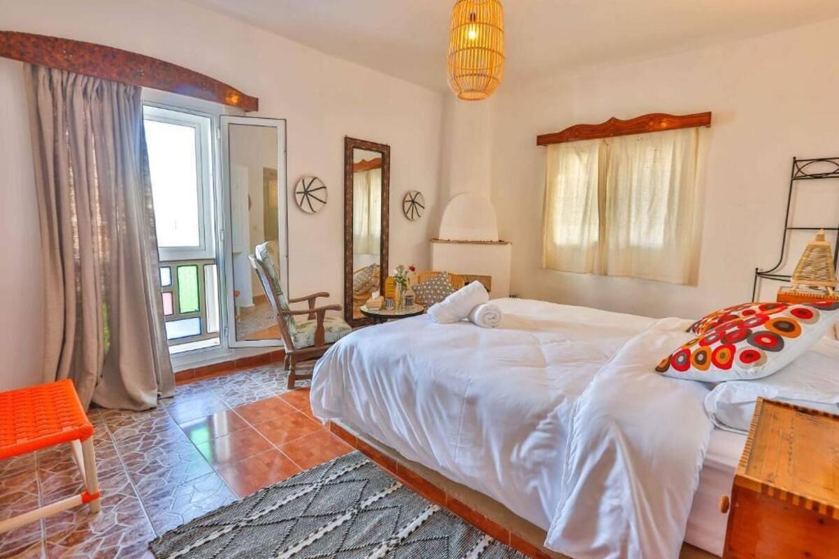 A hotel room with a bed, a chair, a table with a lamp, a window with curtains, and a door leading to a balcony.