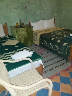A room with two beds, a chair, a nightstand with a lamp, and a patterned rug. - Complexe Collier D'Ambre (Imilchil)