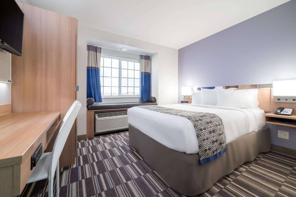 A hotel room with a bed, a desk, a chair, a TV, a window with curtains, and a bedside table with a lamp.