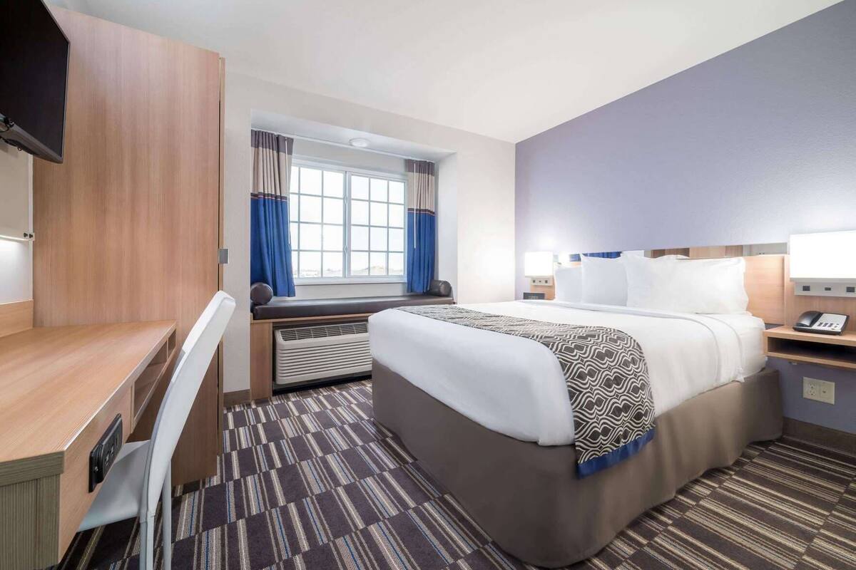 A hotel room with a bed, a desk, a chair, a window with curtains, a heater, and a telephone.