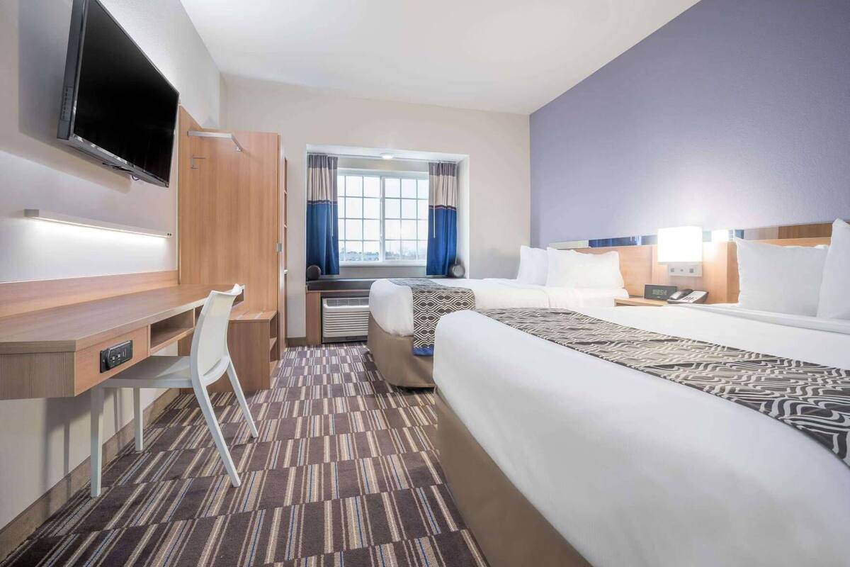 A hotel room with a bed, a desk with a chair, a television, a window with curtains, and a bedside table with a lamp.