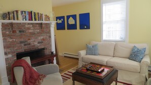 Ein Wohnzimmer mit Kamin aus Ziegelsteinen, einem weißen Sofa mit gemusterten Kissen, einem Couchtisch mit einem Gesellschaftsspiel und einem Bücherregal voller Bücher.