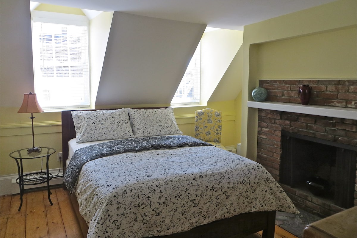 A bedroom with a bed, a small table, a chair, a fireplace, and a window with blinds.