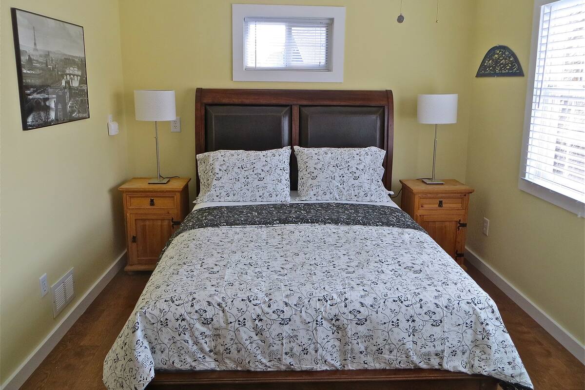 A bedroom with a bed, two nightstands, a window with blinds, a picture on the wall, and a ceiling light.