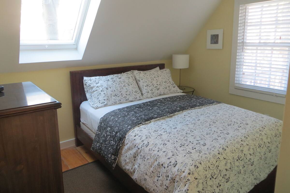 A bedroom with a bed, a desk, a lamp, a window with blinds, and a skylight.