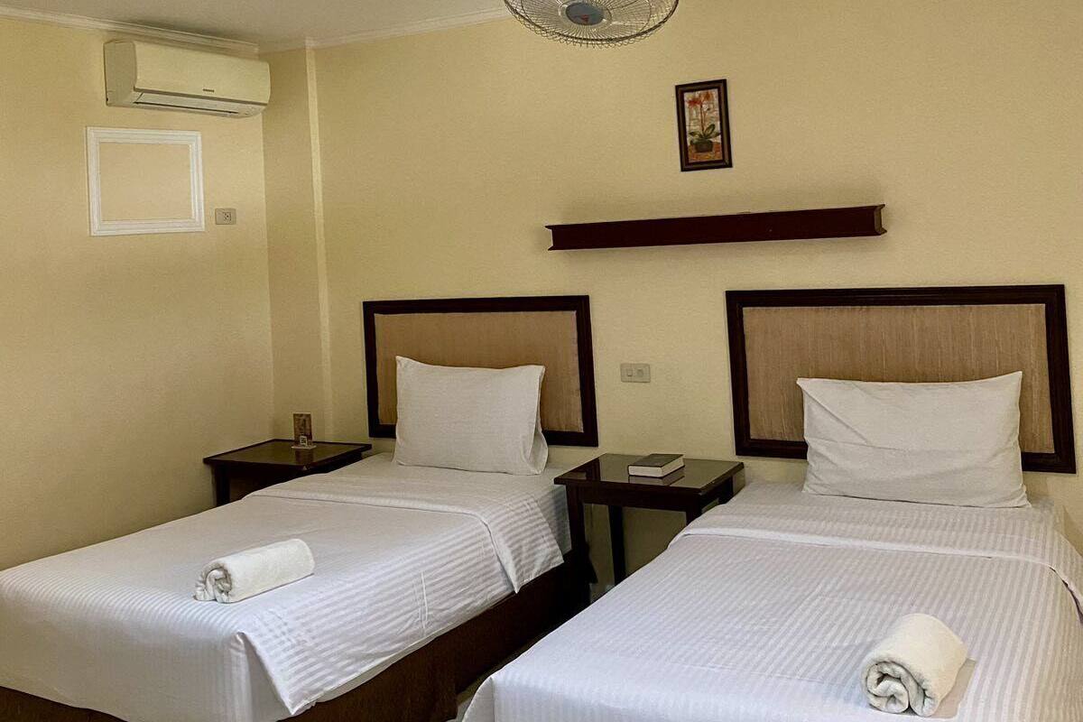 A hotel room with two beds, a ceiling fan, an air conditioning unit, a bedside table with a book, and a framed picture on the wall.
