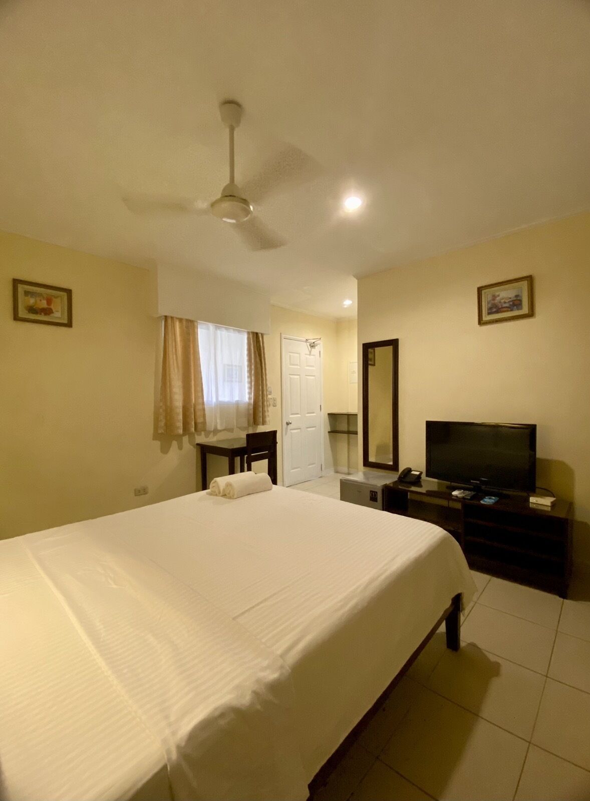 A hotel room with a bed, a desk, a television, a mirror, and a ceiling fan.