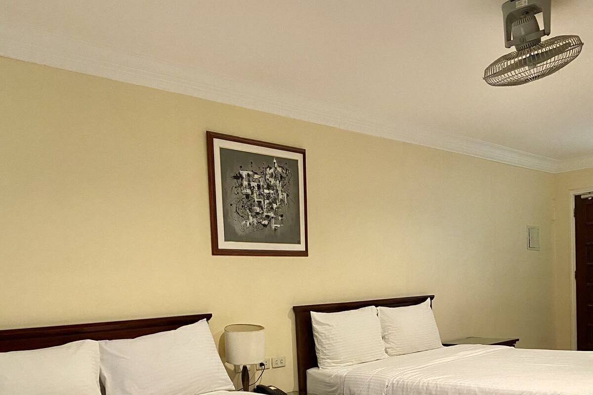 A hotel room with two beds, a nightstand with a lamp, a ceiling fan, and a framed picture on the wall.