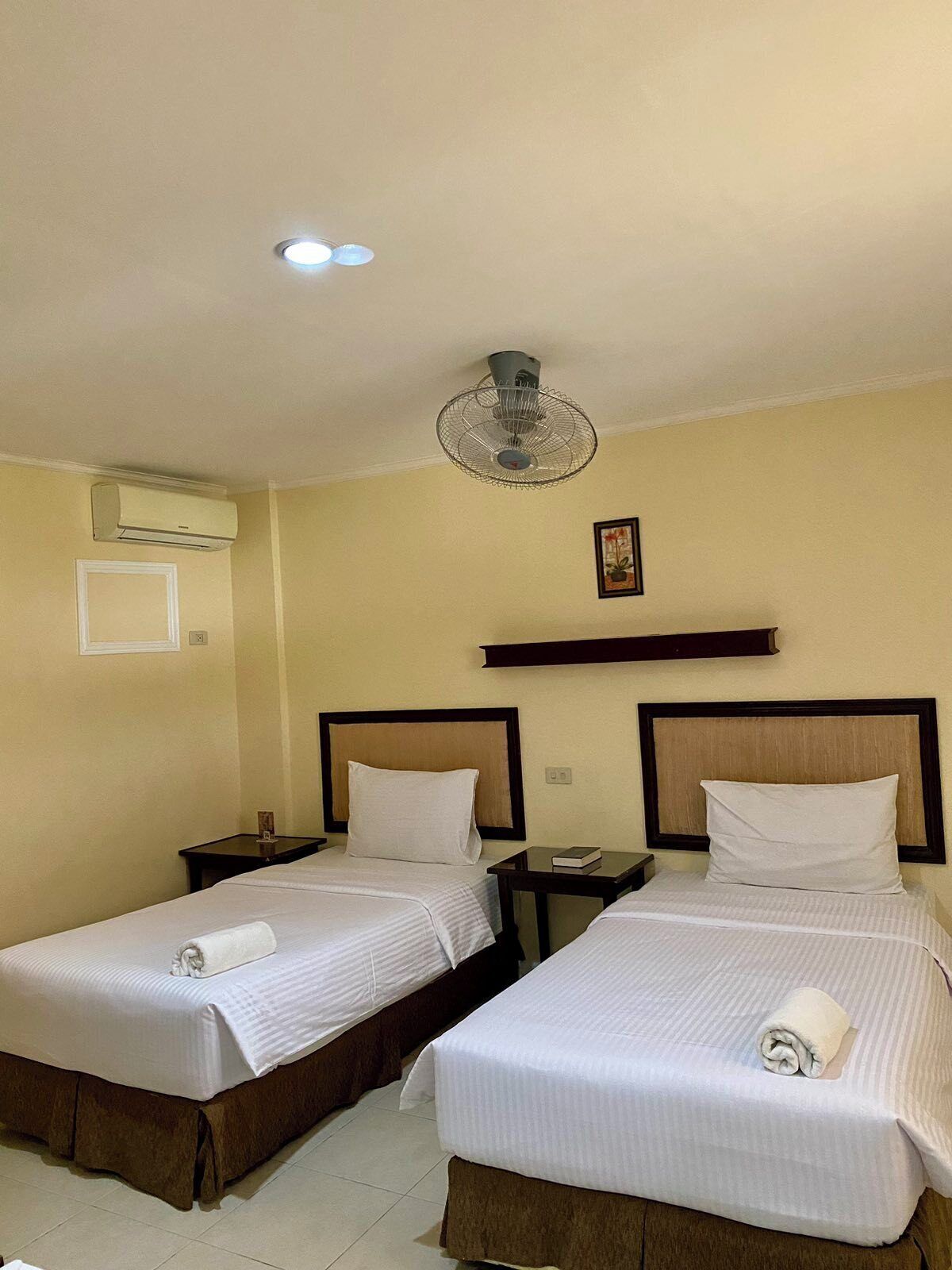 Ein Hotelzimmer mit zwei Betten, einem Deckenventilator, einer Klimaanlage, einem Nachttisch mit einem Buch und einem gerahmten Bild an der Wand.
