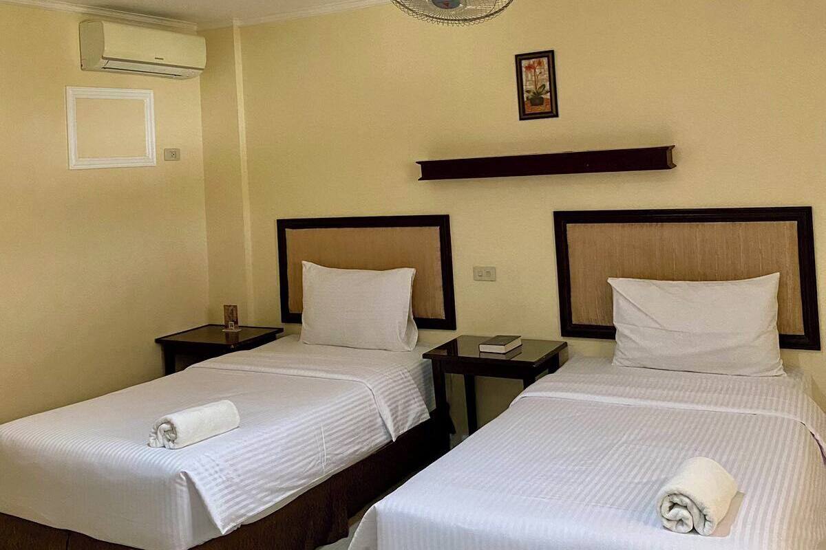 Een hotelkamer met twee bedden, een plafondventilator, een airconditioner, een nachtkastje met een boek en een ingelijste foto aan de muur.