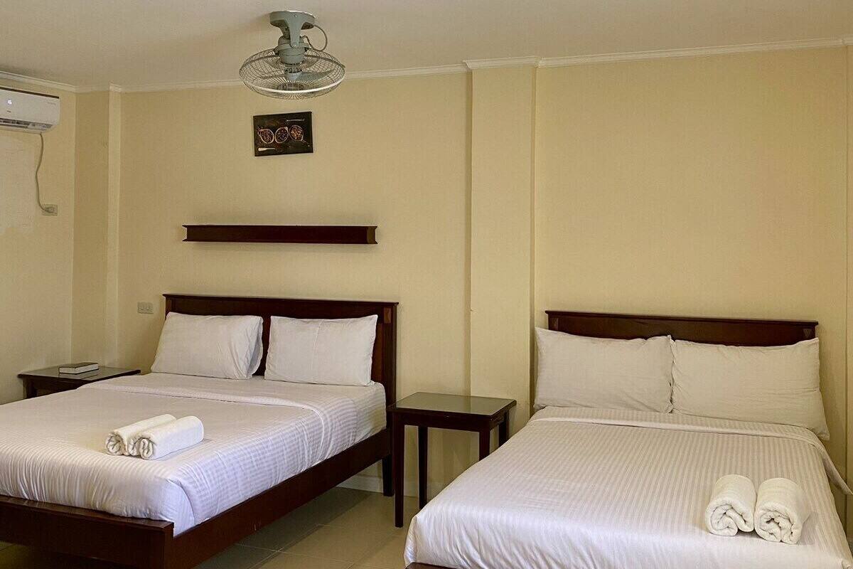 A hotel room with two single beds, a ceiling fan, a small table, and an air conditioning unit.