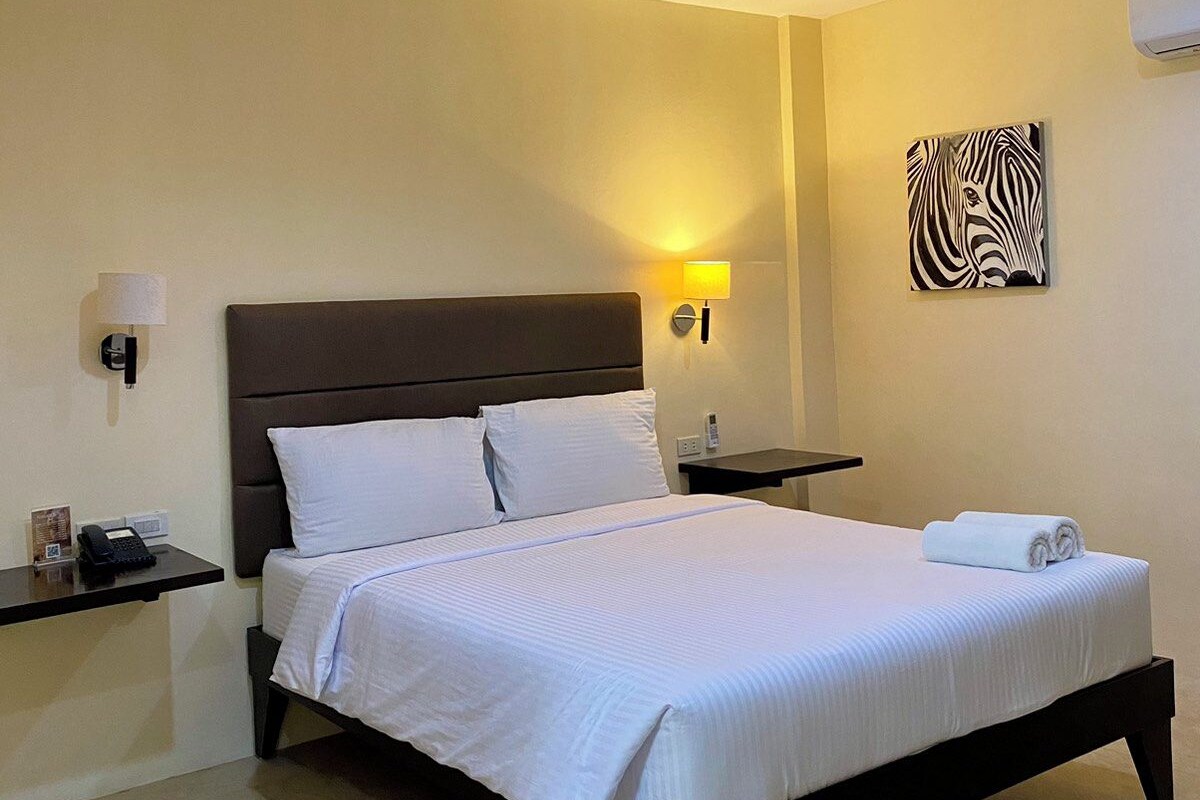 Une chambre d’hôtel avec un grand lit, un ventilateur de plafond, un climatiseur mural, une lampe de chevet et une œuvre d’art représentant un zèbre accrochée au mur.