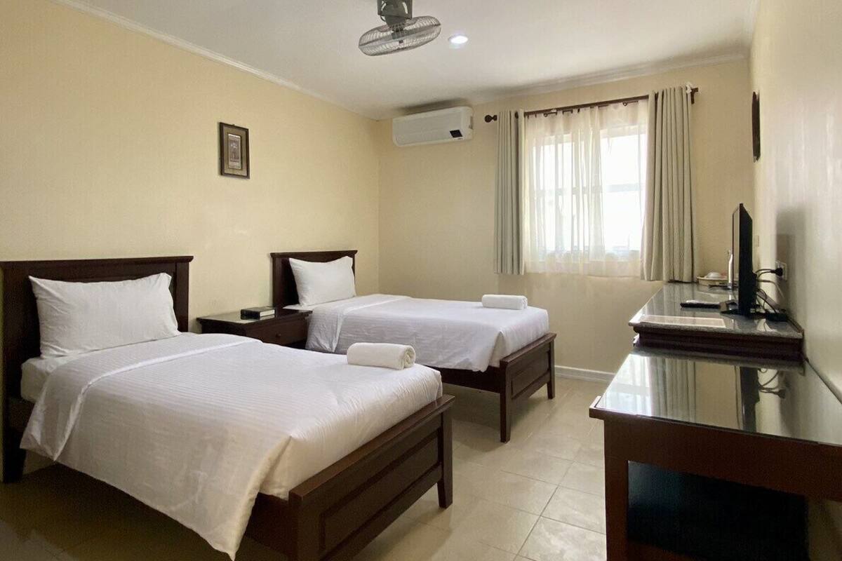 A hotel room with two single beds, a desk, a chair, a ceiling fan, an air conditioning unit, and a window with curtains.