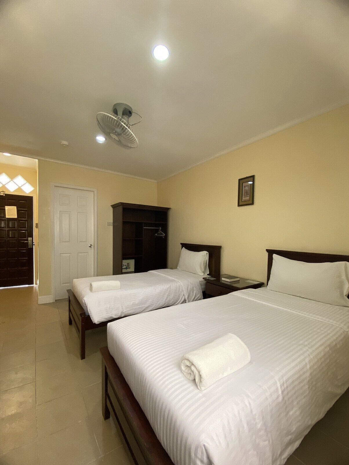 A hotel room with two beds, a wardrobe, a bedside table, a ceiling fan, and a door leading to a hallway.