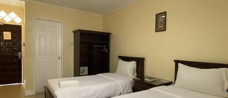 Habitación de hotel con dos camas, un armario, una mesita de noche, un ventilador de techo y una puerta que da a un pasillo.