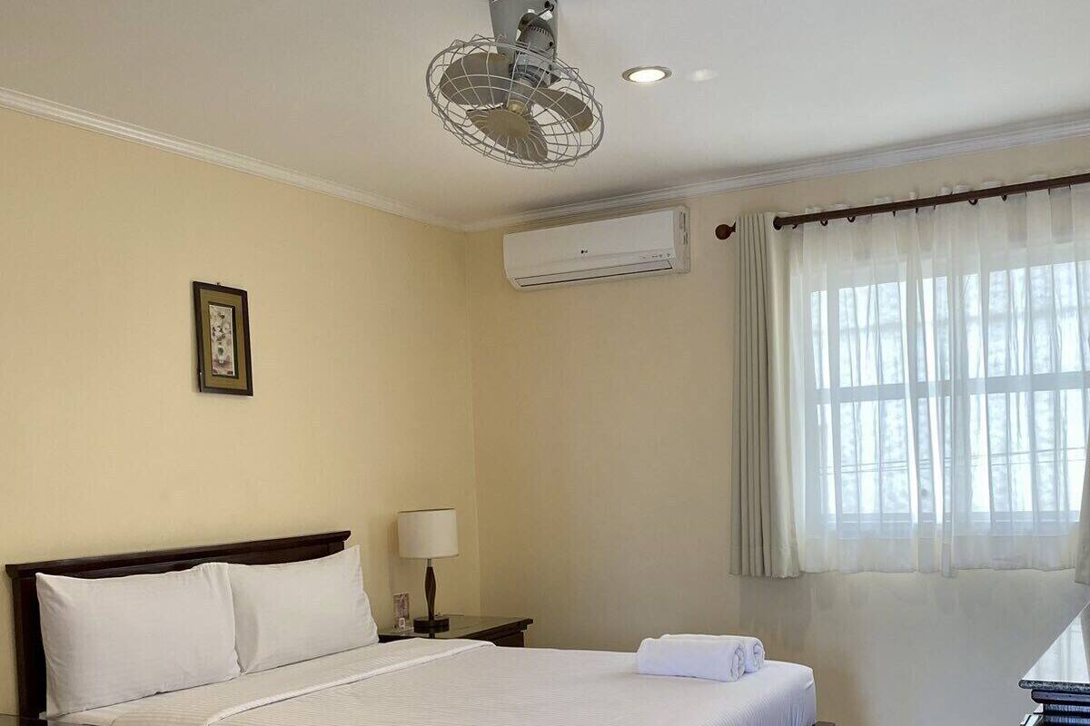A hotel room with a bed, bedside table, lamp, air conditioning unit, and a window with curtains.