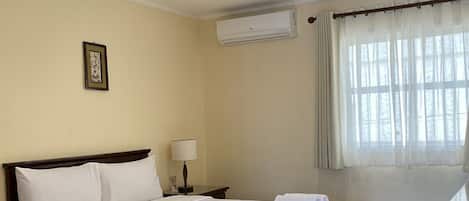 A hotel room with a bed, bedside table, lamp, air conditioning unit, and a window with curtains.