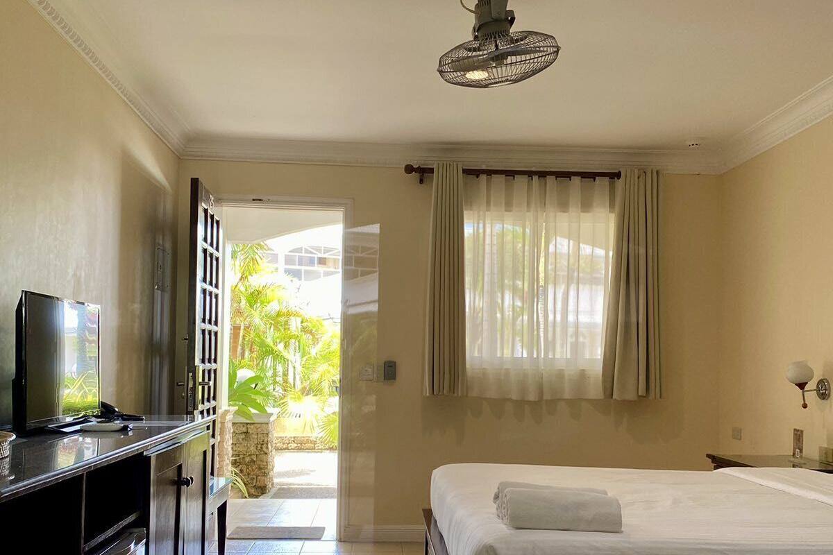 A hotel room with a bed, a desk with a TV, a ceiling fan, and a view of the outdoors.