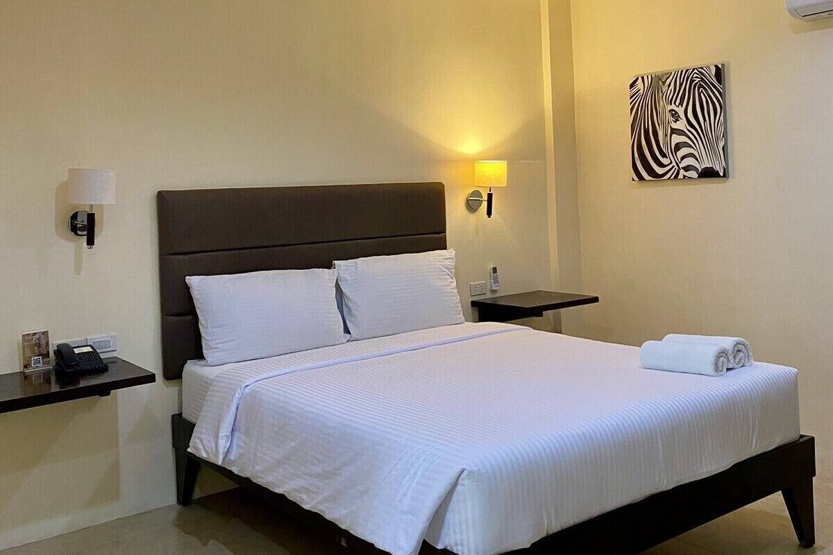 A hotel room with a large bed, a ceiling fan, a wall-mounted air conditioner, a bedside lamp, and a zebra artwork on the wall.