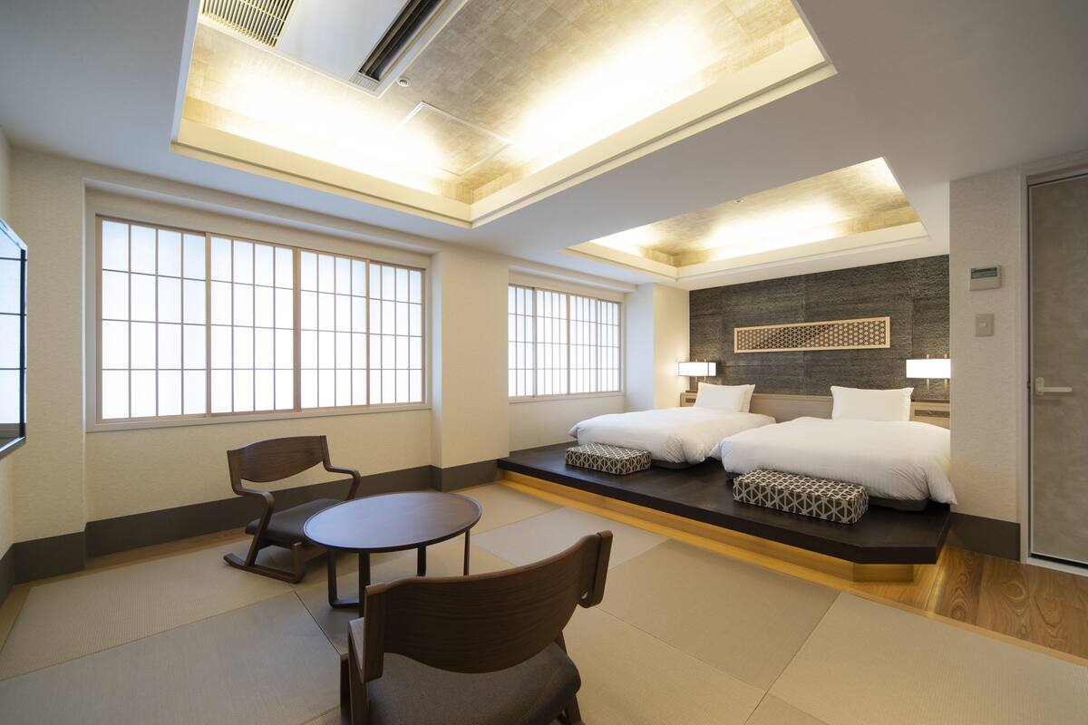 Habitación de hotel con cama, dos sillas, una mesita y ventanas amplias.