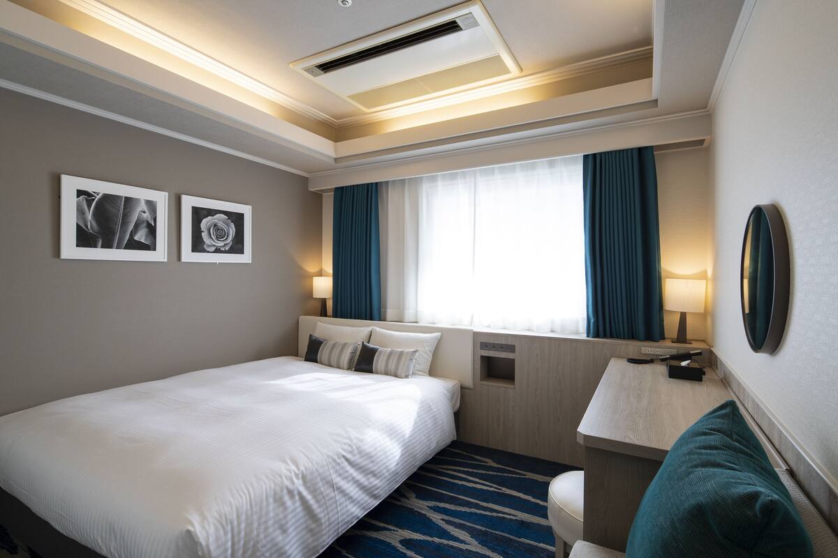 Habitación de hotel con cama, escritorio, silla, ventana con cortinas y dos cuadros en la pared.