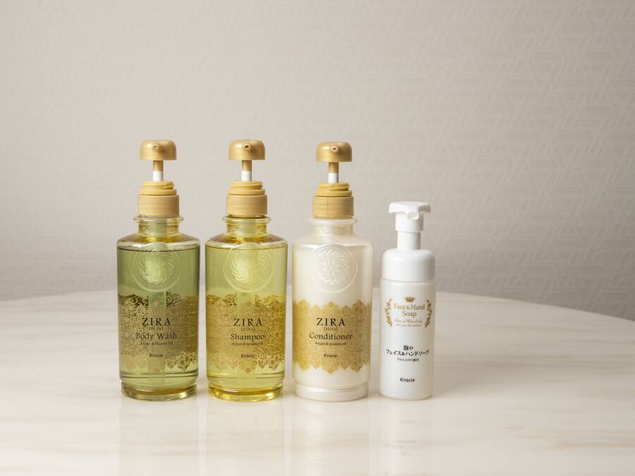 Cuatro botellas de jabón de baño, champú, acondicionador y jabón de manos ZIRA dispuestas sobre una superficie blanca.
