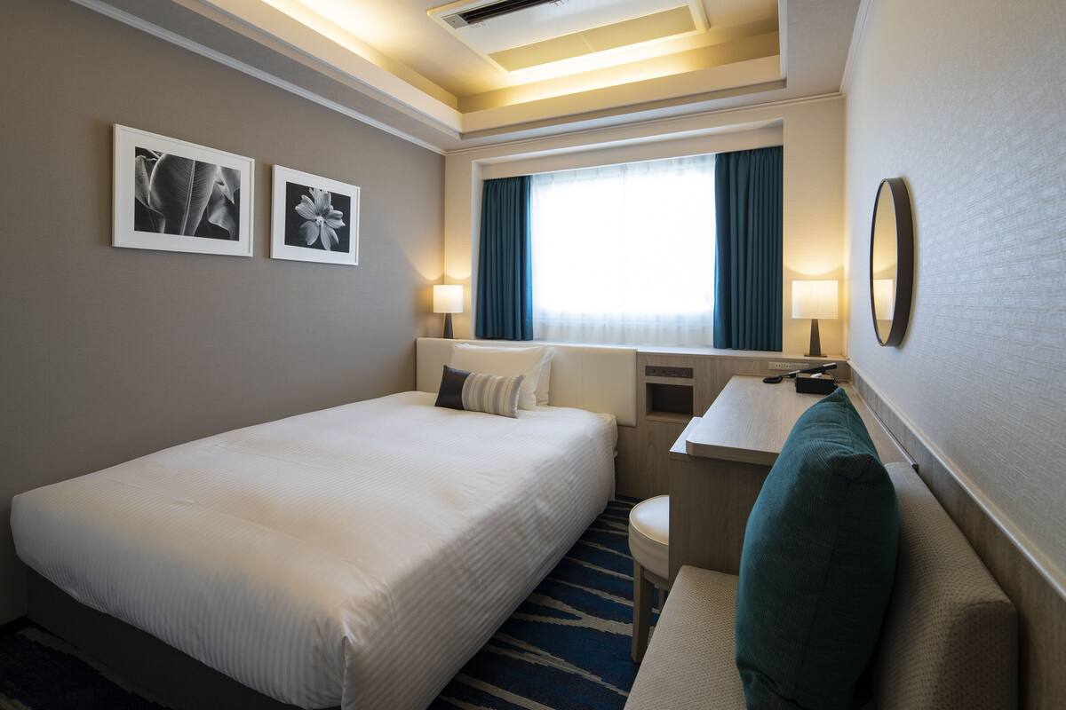 Habitación de hotel con una cama grande, un escritorio, una silla, un ventanal con cortinas y dos cuadros en la pared.