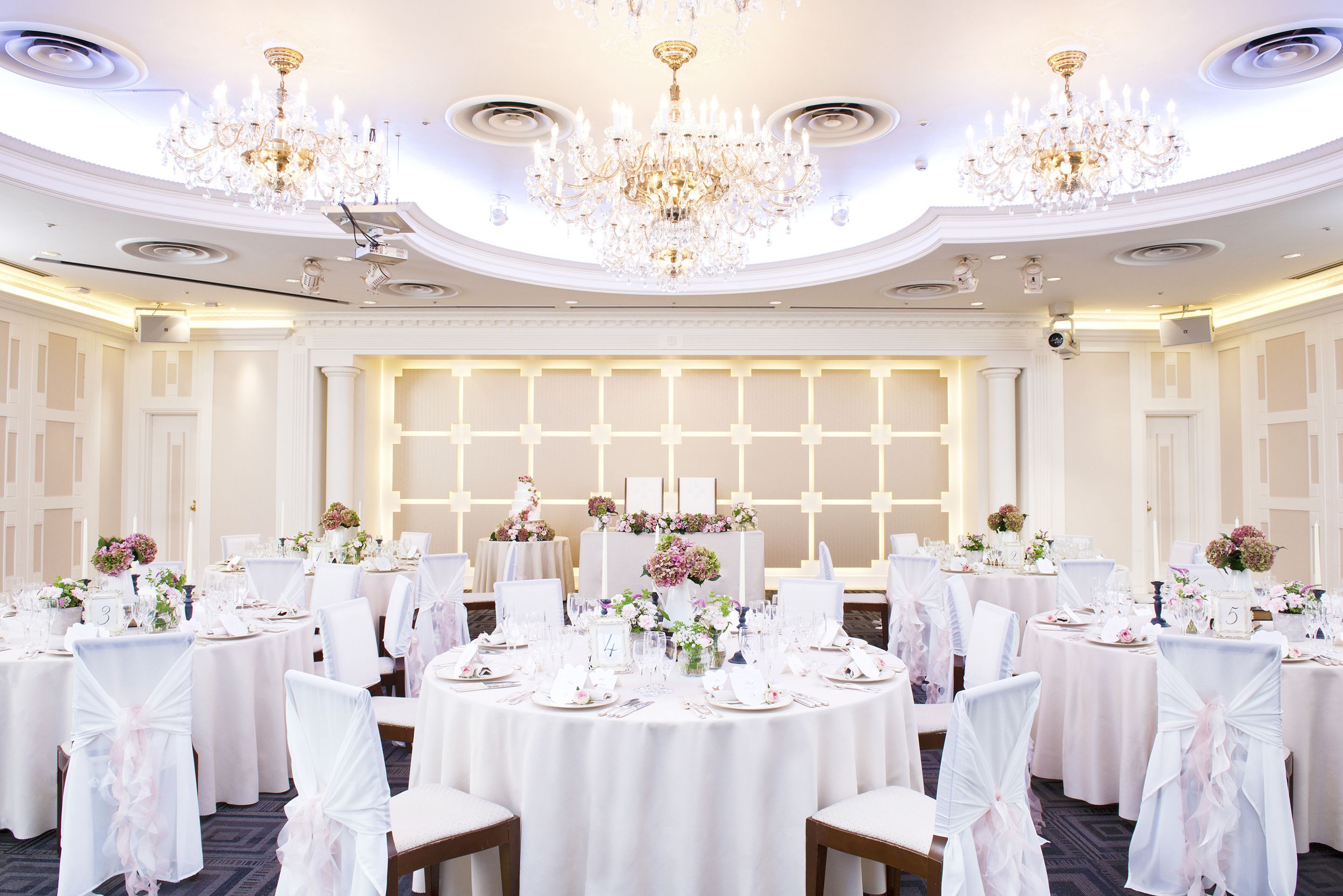 A banquet hall with round tables, white chairs, chandeliers, and floral centerpieces.