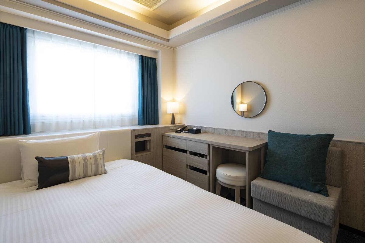 Habitación de hotel con cama, escritorio, silla, espejo redondo y ventana con cortinas.