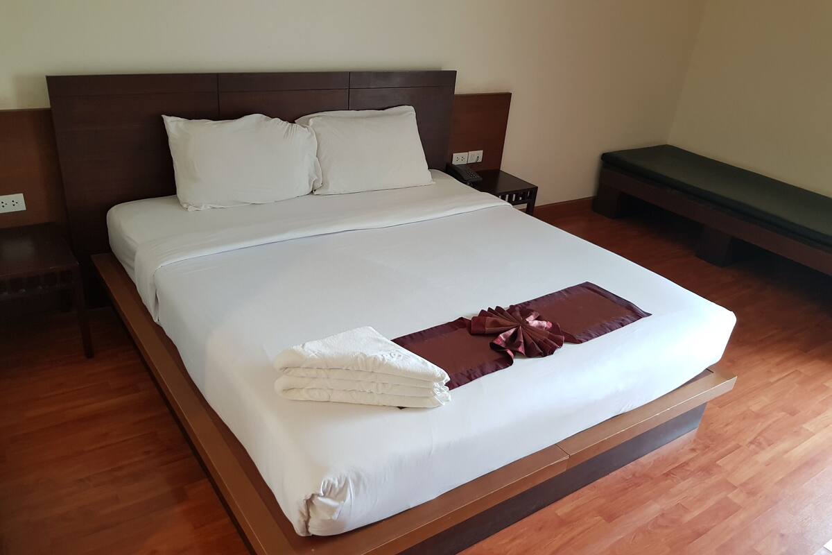 A neatly made bed with white linens and a decorative bow, a wooden headboard, a bedside table with a phone, and a bench against the wall.