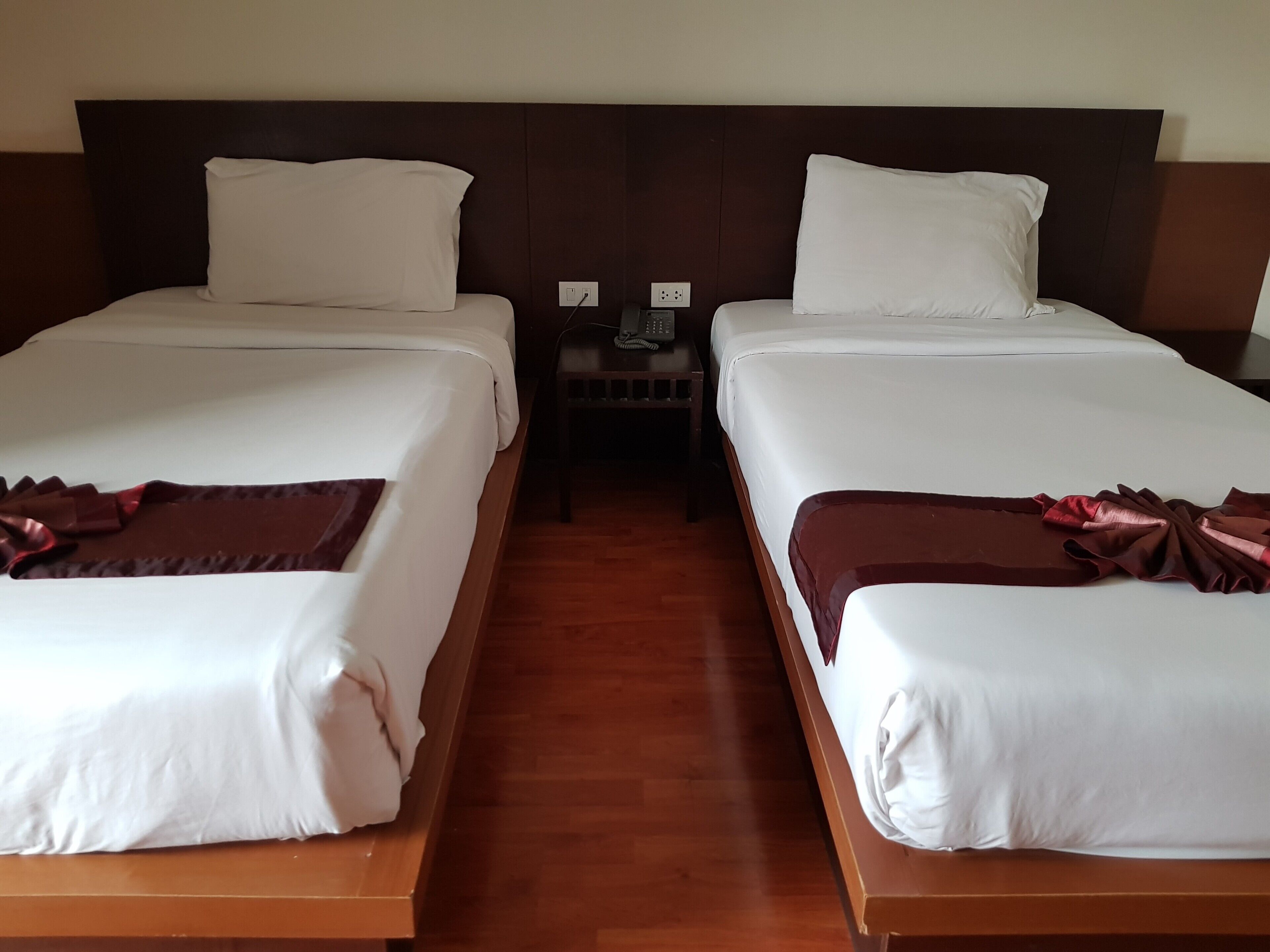 Two single beds with white linens and brown decorative pillows, a wooden headboard, a nightstand with a phone, and a power outlet.