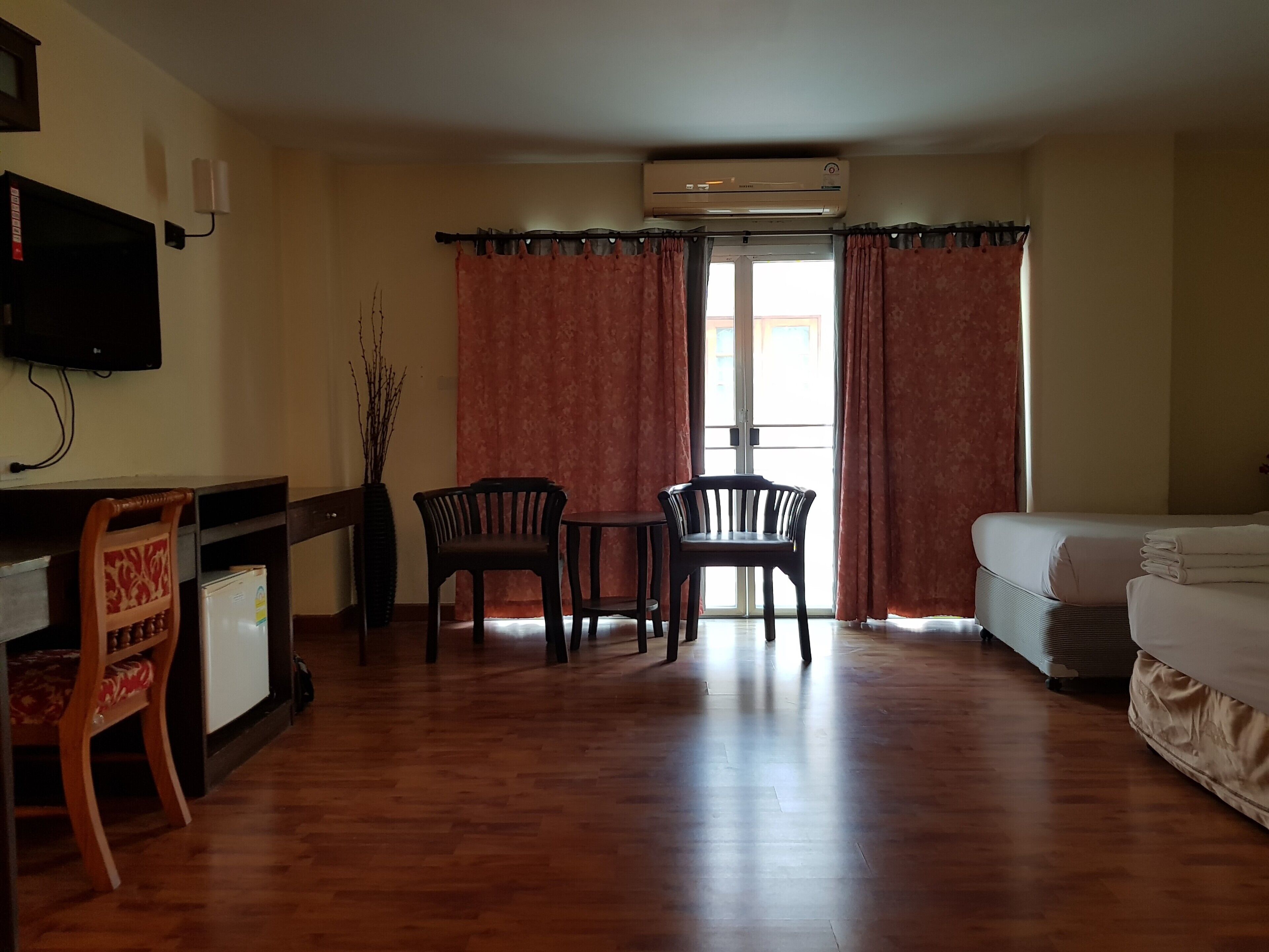 Habitación de hotel con suelo de madera, una cama, un escritorio con silla, una pequeña mesa con dos sillas, un equipo de aire acondicionado y un televisor en la pared.