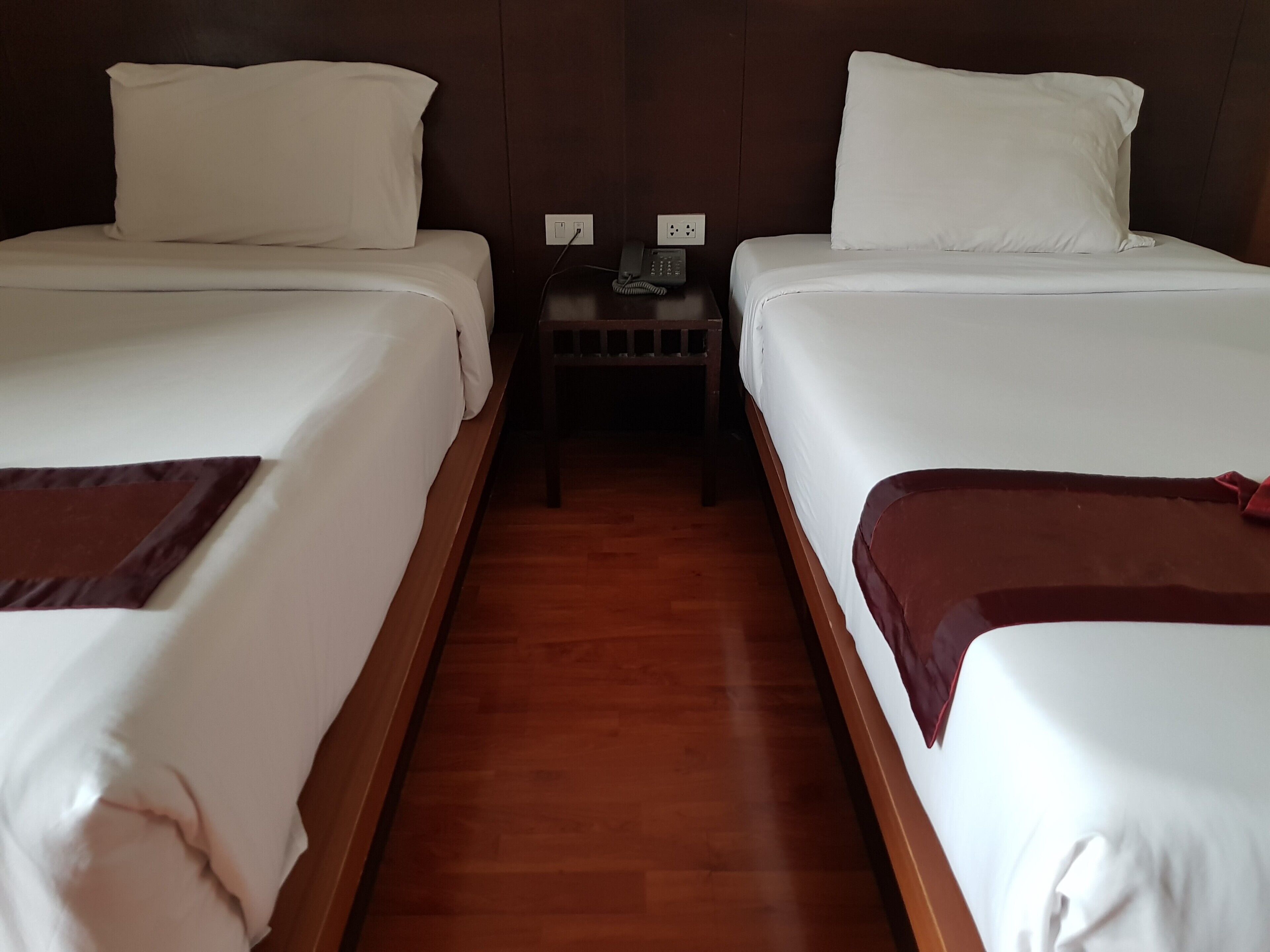 A hotel room with two single beds, wooden flooring, a nightstand with a telephone, and a wall socket.