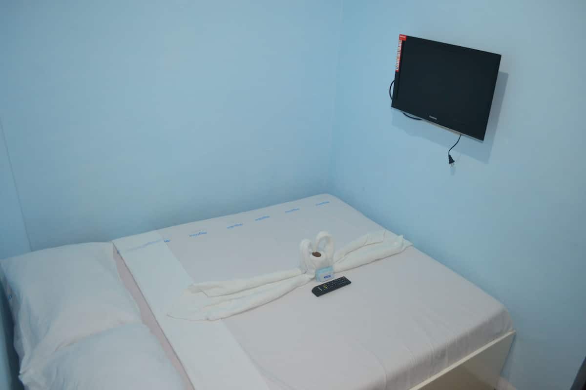 Una cama individual con una colcha blanca y un control remoto encima. Hay un televisor de pantalla plana montado en la pared sobre la cama.