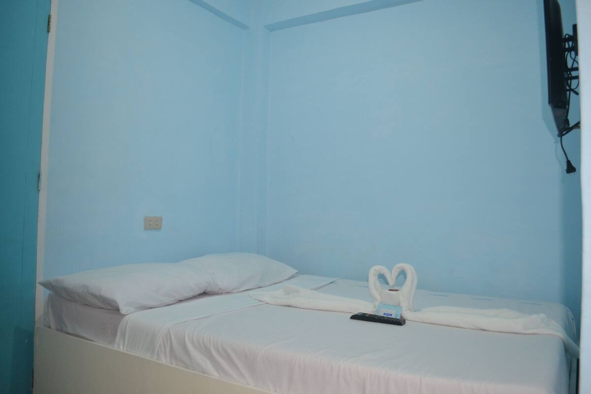Una cama individual con ropa blanca, un control remoto y una toalla con forma de cisne sobre una pared azul.