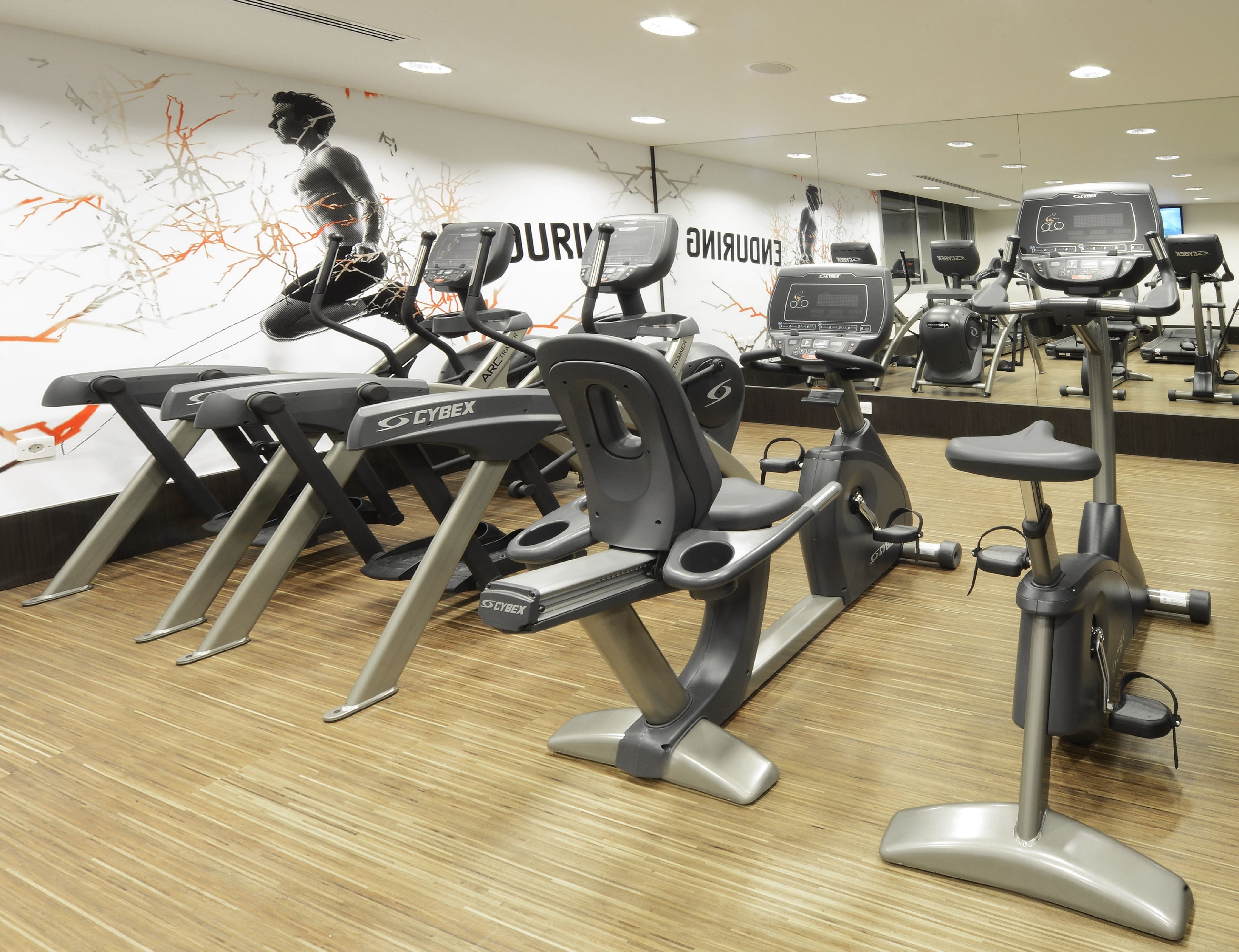 Un gimnasio con varias bicicletas estáticas y cintas de correr, suelo de madera, y una pared con arte abstracto y la palabra “DURING”. 