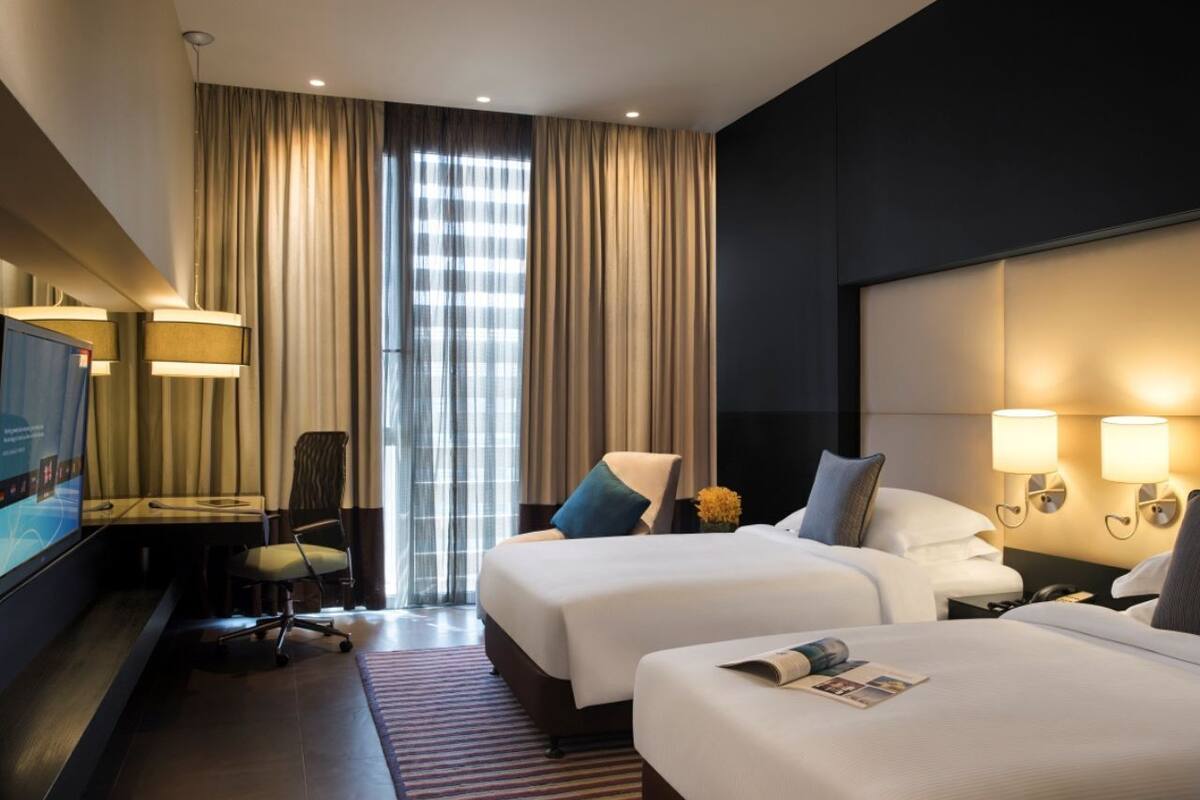 Quarto de hotel com duas camas, uma escrivaninha com cadeira, televisão e uma janela grande com cortinas.