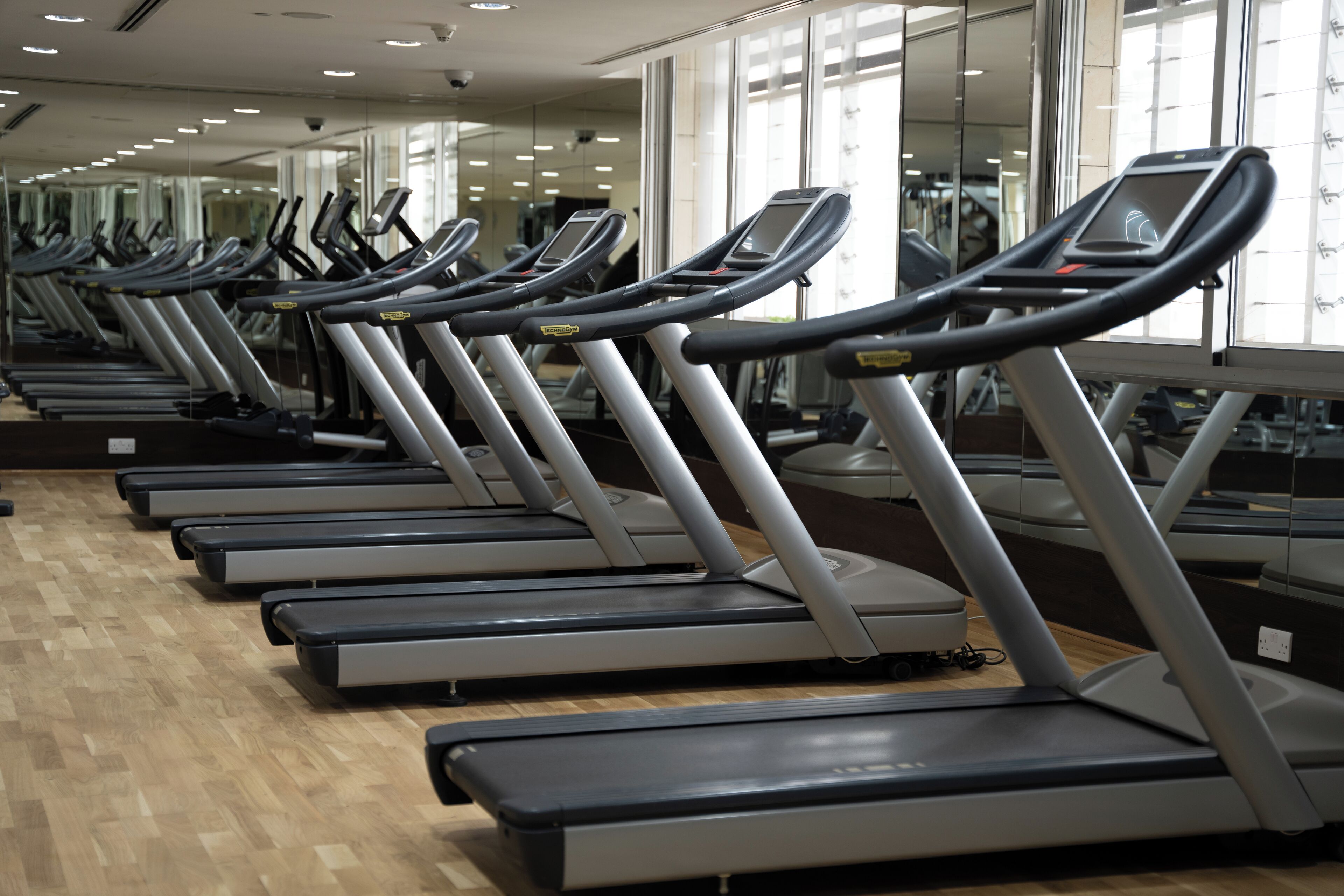 A row of stationary treadmills in a gym with large windows and a wooden floor.