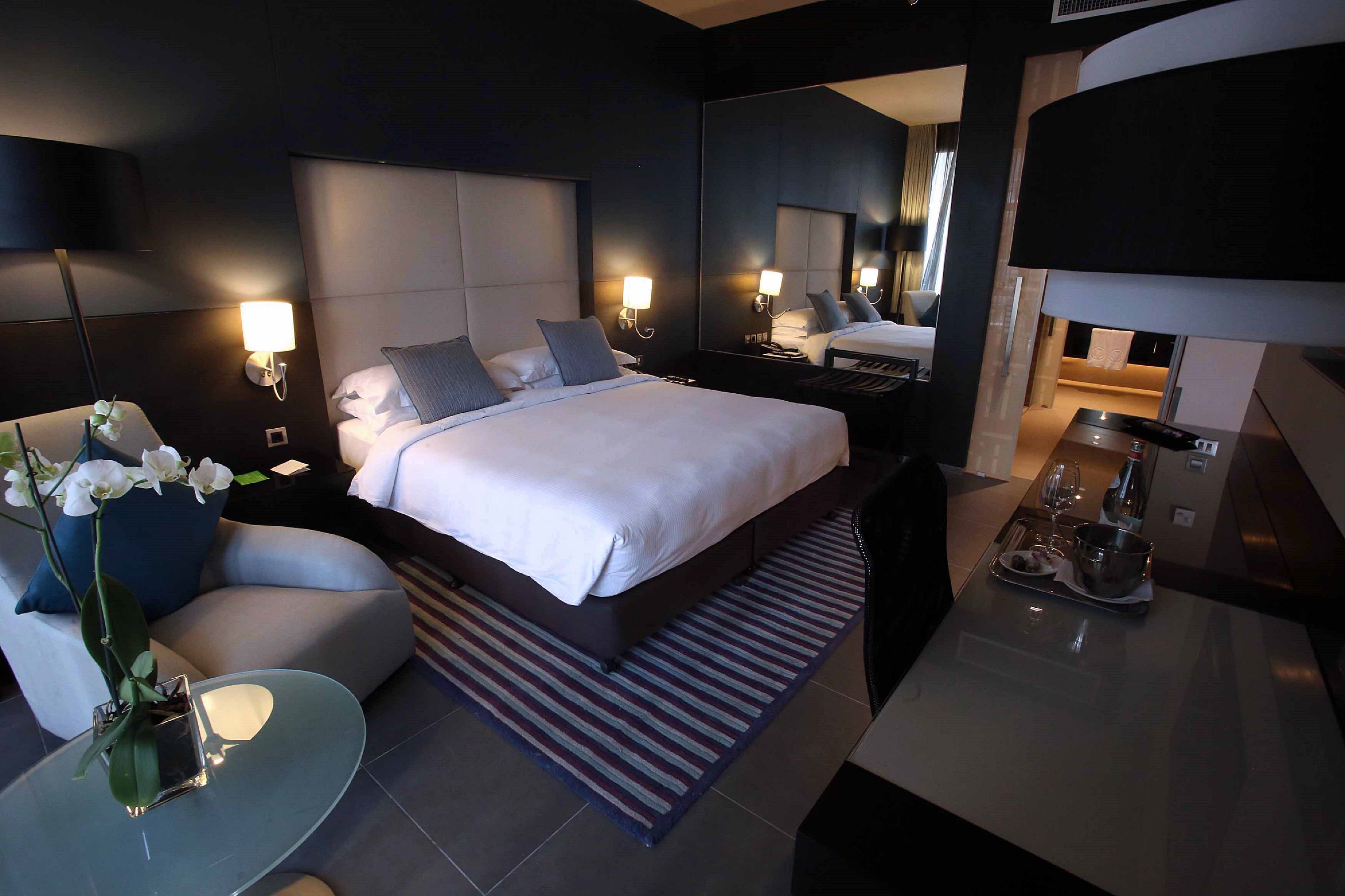 A modern hotel room with a large bed, bedside tables, a small seating area, and a dining table with a tray of items.