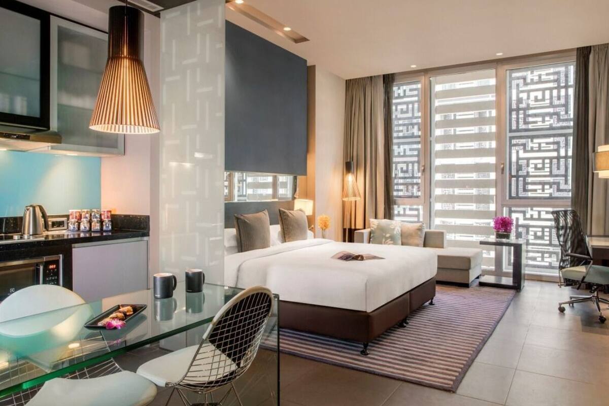Quarto de hotel moderno com uma cama grande, uma mesa de jantar de vidro, uma área de cozinha e vista pela janela com padrão.