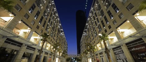 A modern urban street with high-rise buildings, palm trees, and illuminated signs.