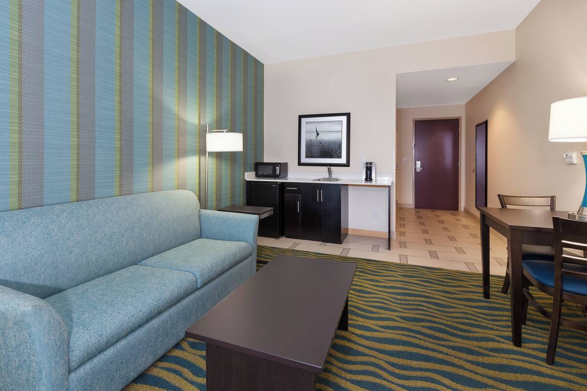 Habitación de hotel con un sofá azul, una mesita pequeña, una zona de cocina con microondas, cafetera y un cuadro en la pared.