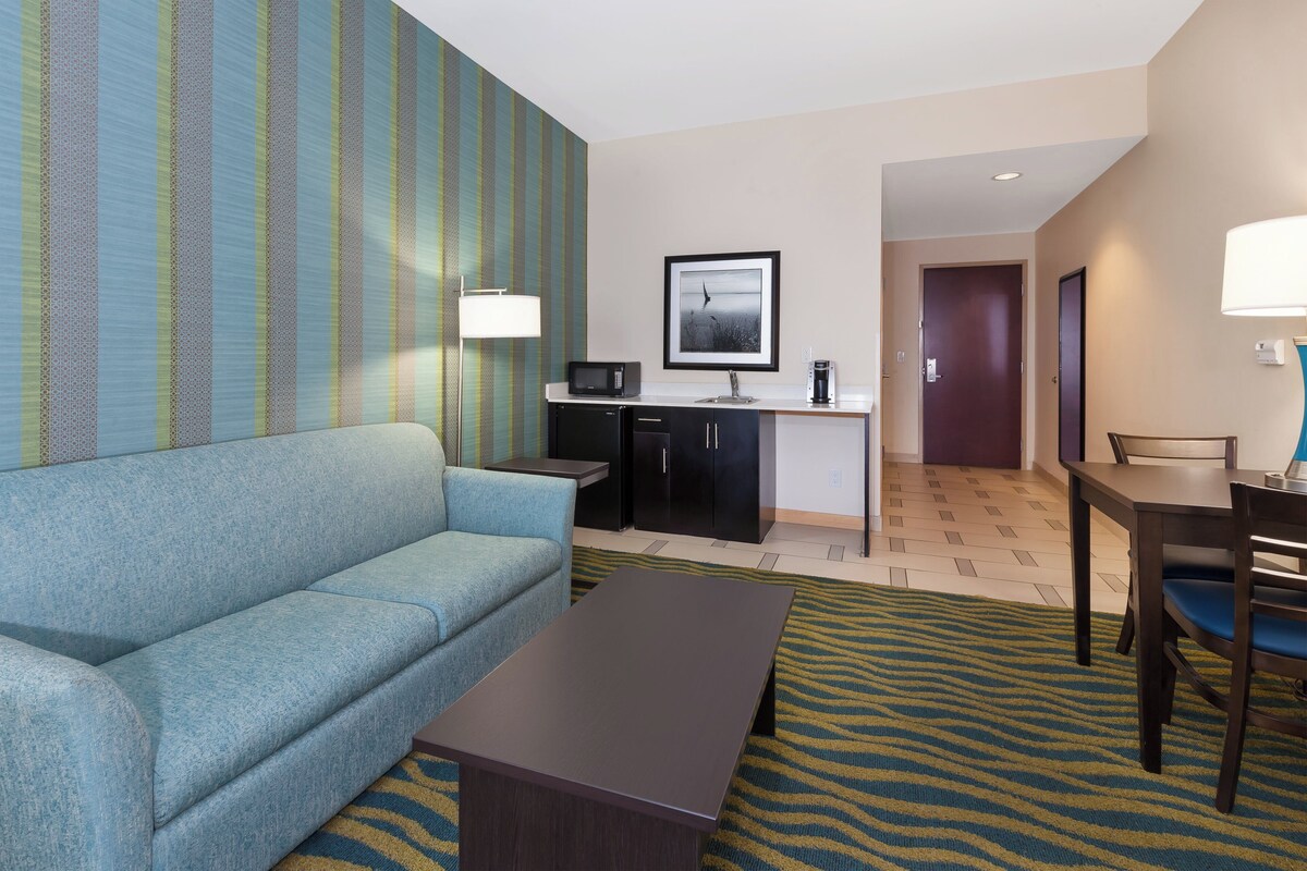 Een hotelkamer met een blauwe bank, een kleine tafel, een kitchenette met een magnetron, een koffiezetapparaat en een lijst aan de muur.