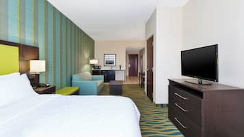 Quarto de hotel com uma cama grande, televisão, escrivaninha com luminária, cadeira e uma pequena cozinha.