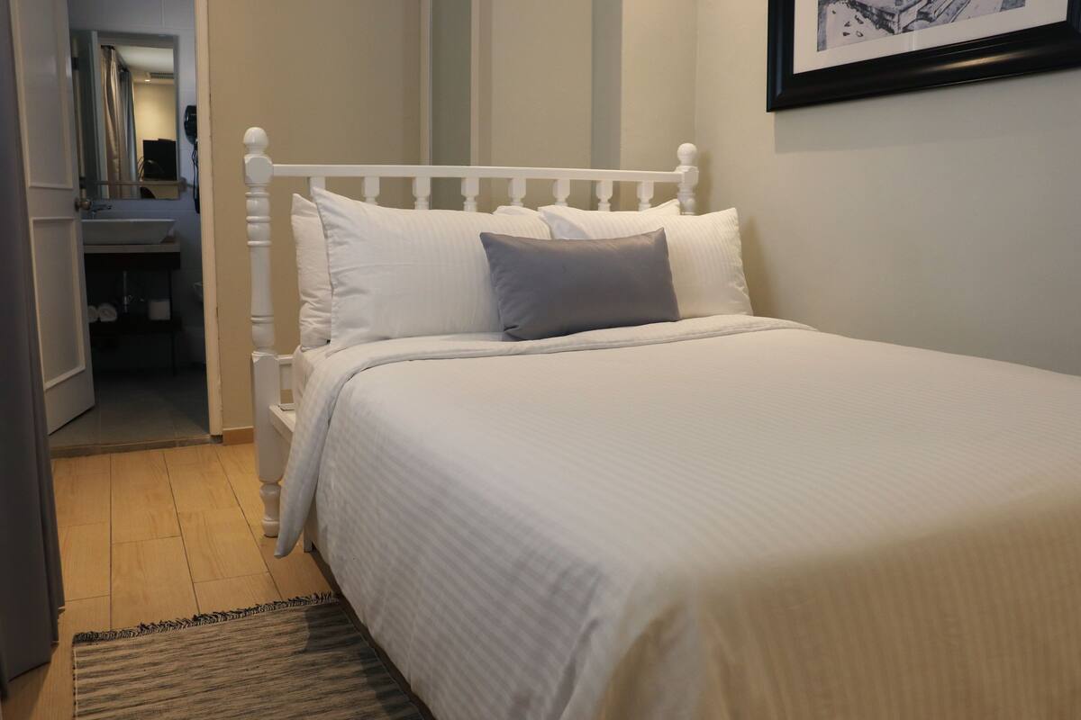 A neatly made bed with white bedding and a gray pillow in a room with a wooden floor and a small rug.