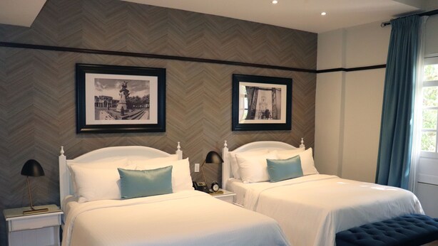 Habitación de hotel con dos camas, un banco, una mesita de noche con lámpara y cuadros en la pared.