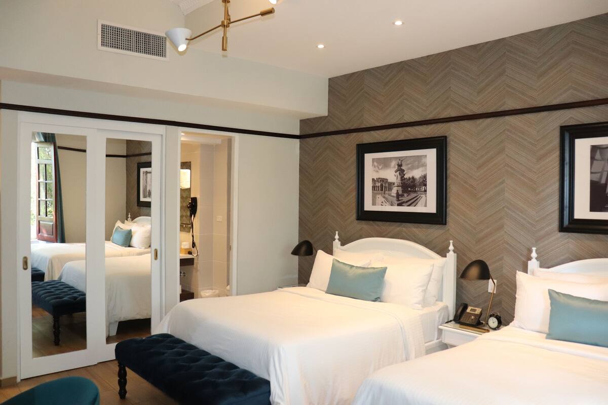 A hotel room with two beds, a mirror, a bench, and a nightstand with a lamp and phone.