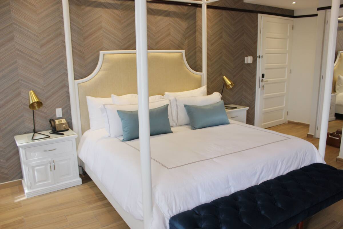 A four-poster bed with a white headboard, bedside tables with lamps, a bench, and a herringbone-patterned wall.